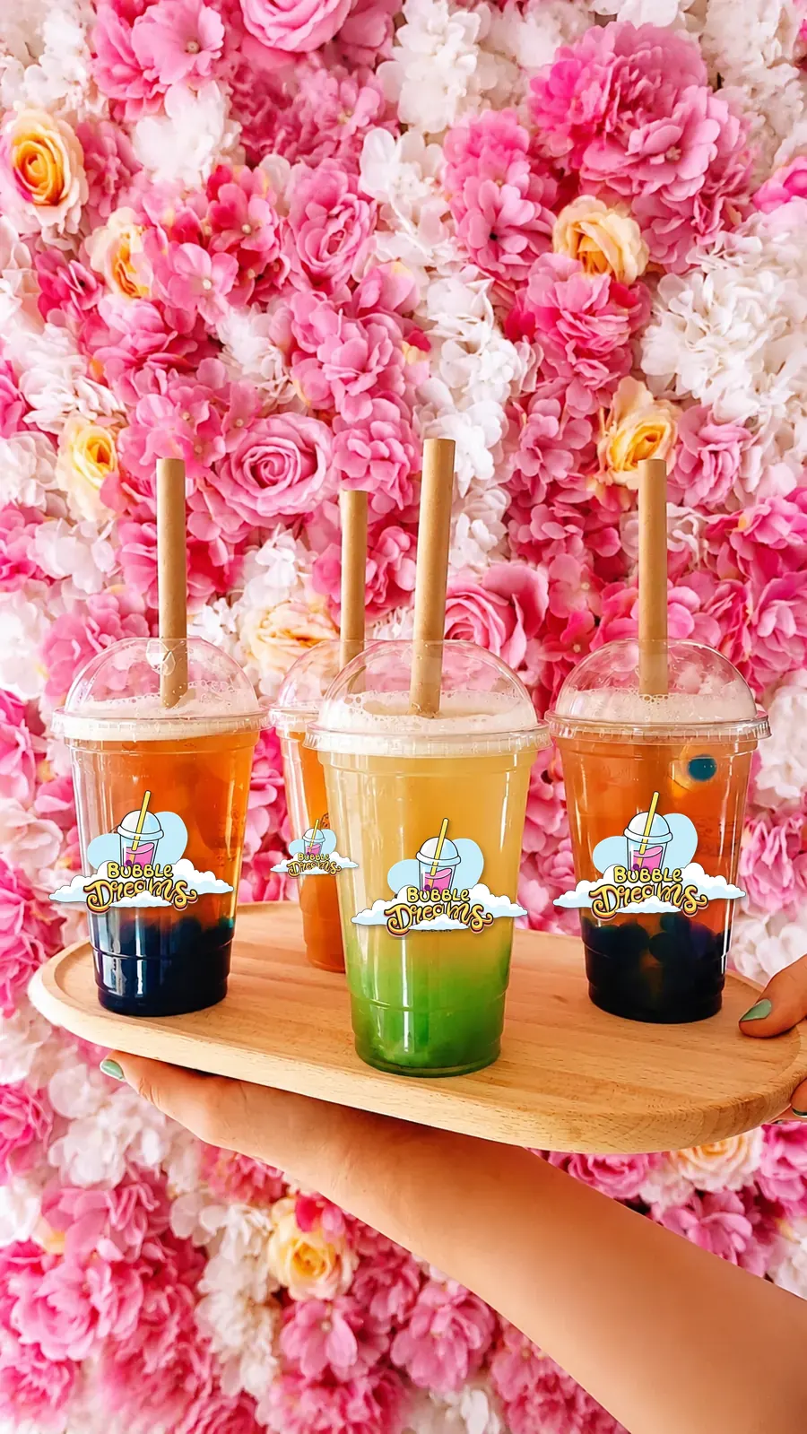 Bandeja con tres bubble teas de colores y perlas de fruta sobre un muro de flores rosas